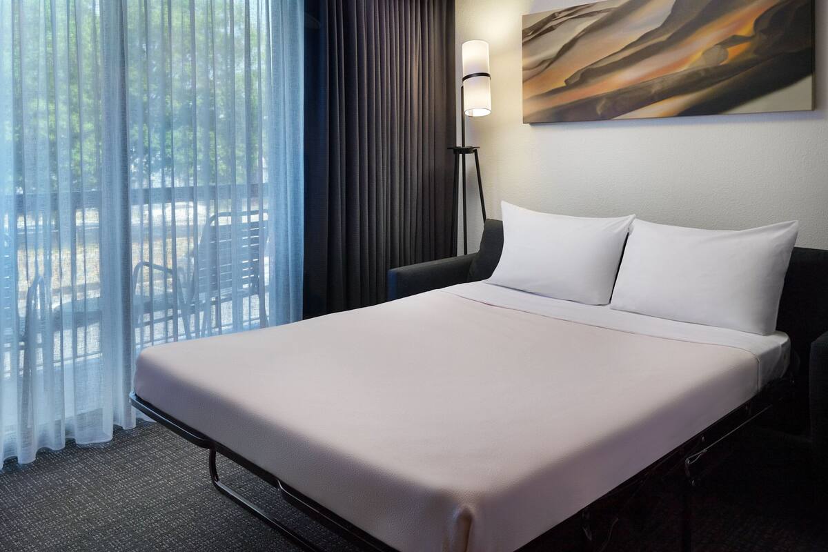 A hotel room with a large bed, a standing lamp, a painting on the wall, and a view through sheer curtains.