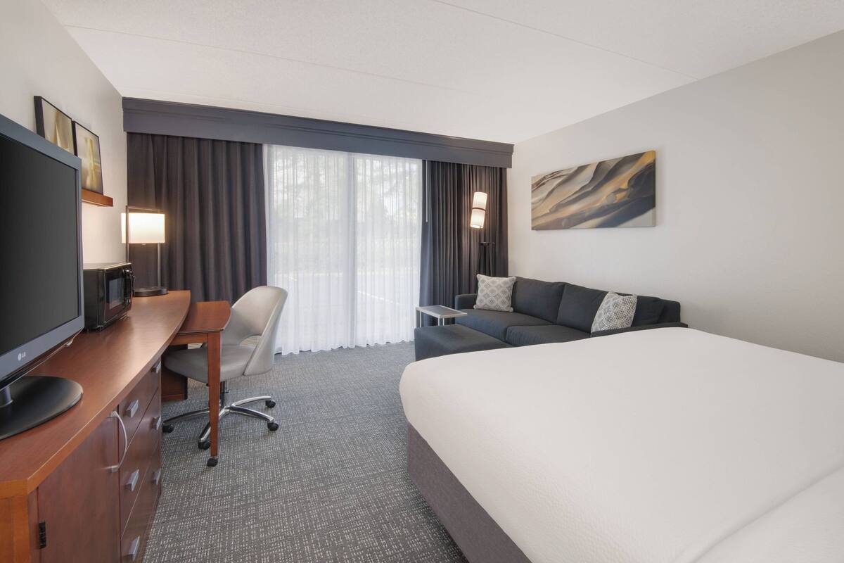 A hotel room with a bed, a desk with a TV, a chair, a sofa, and a large window with curtains.