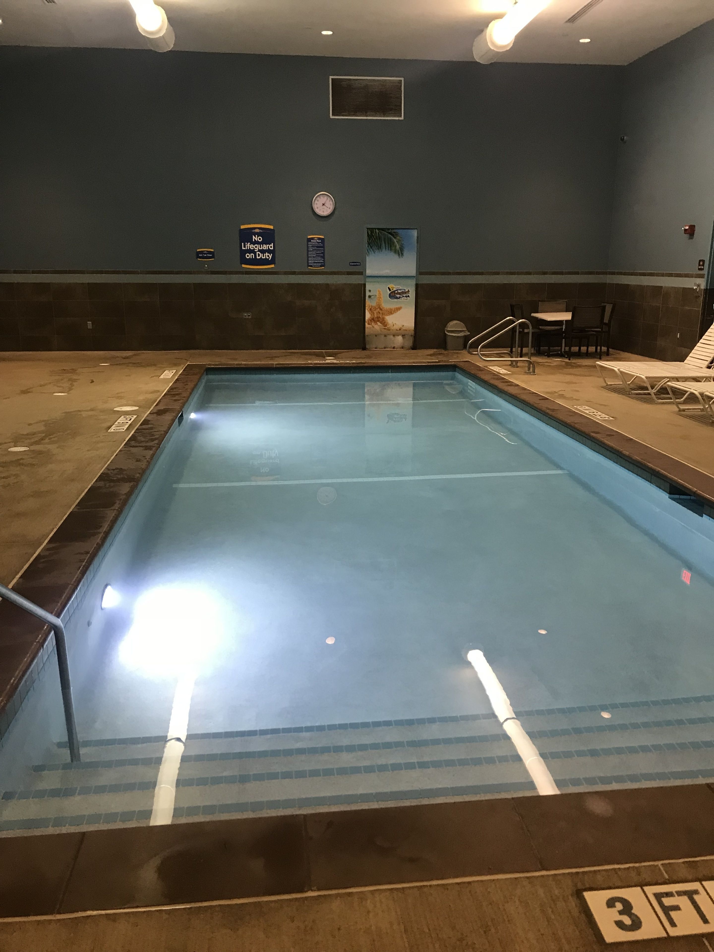 An indoor swimming pool with a 3 FT depth marking, lounge chairs, and a lifeguard duty sign.