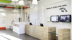 A modern reception area with a wooden desk, a glass partition, and a wall-mounted TV displaying a show. - ibis Styles Nancy Sud (Houdemont)