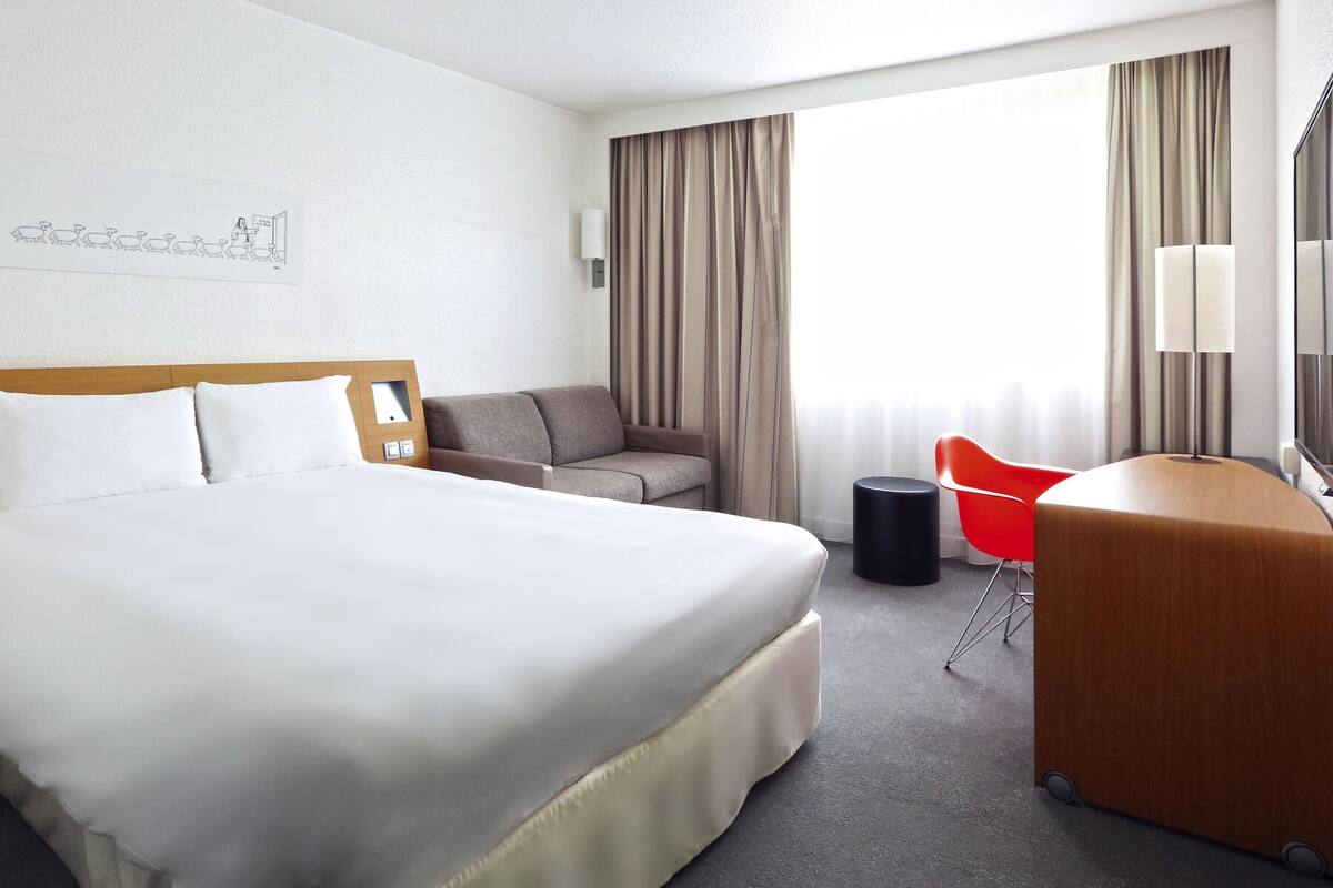 A hotel room with a large bed, a sofa, a desk with a chair, a lamp, and a window with curtains.