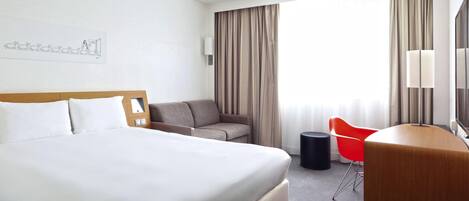 A hotel room with a large bed, a sofa, a desk with a chair, a lamp, and a window with curtains.
