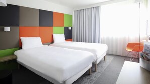 A hotel room with two beds, a desk, a chair, and a large window with curtains. - ibis Styles Nancy Sud (Houdemont)