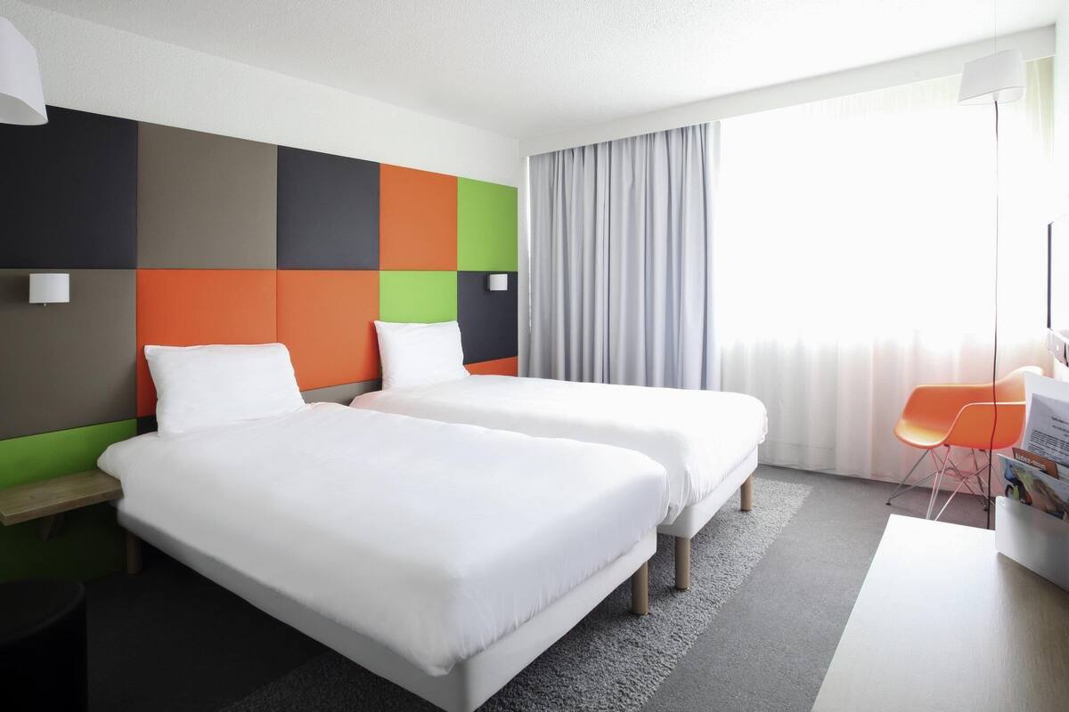 A hotel room with two beds, a desk, a chair, and a large window with curtains.