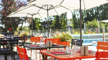 Un espace extérieur aménagé pour les repas, avec des tables, des chaises, des parasols et une piscine en arrière-plan.