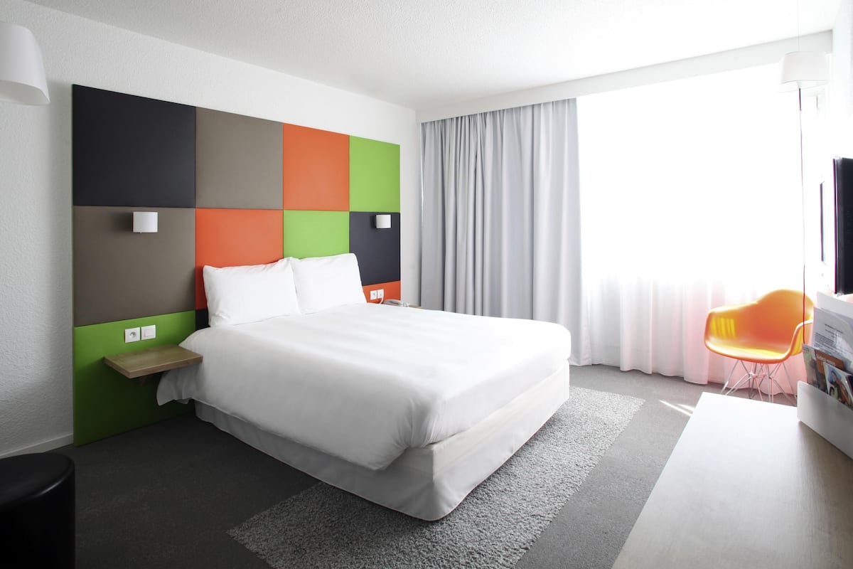 A modern hotel room with a large bed, a bedside table, a chair, and a TV.
