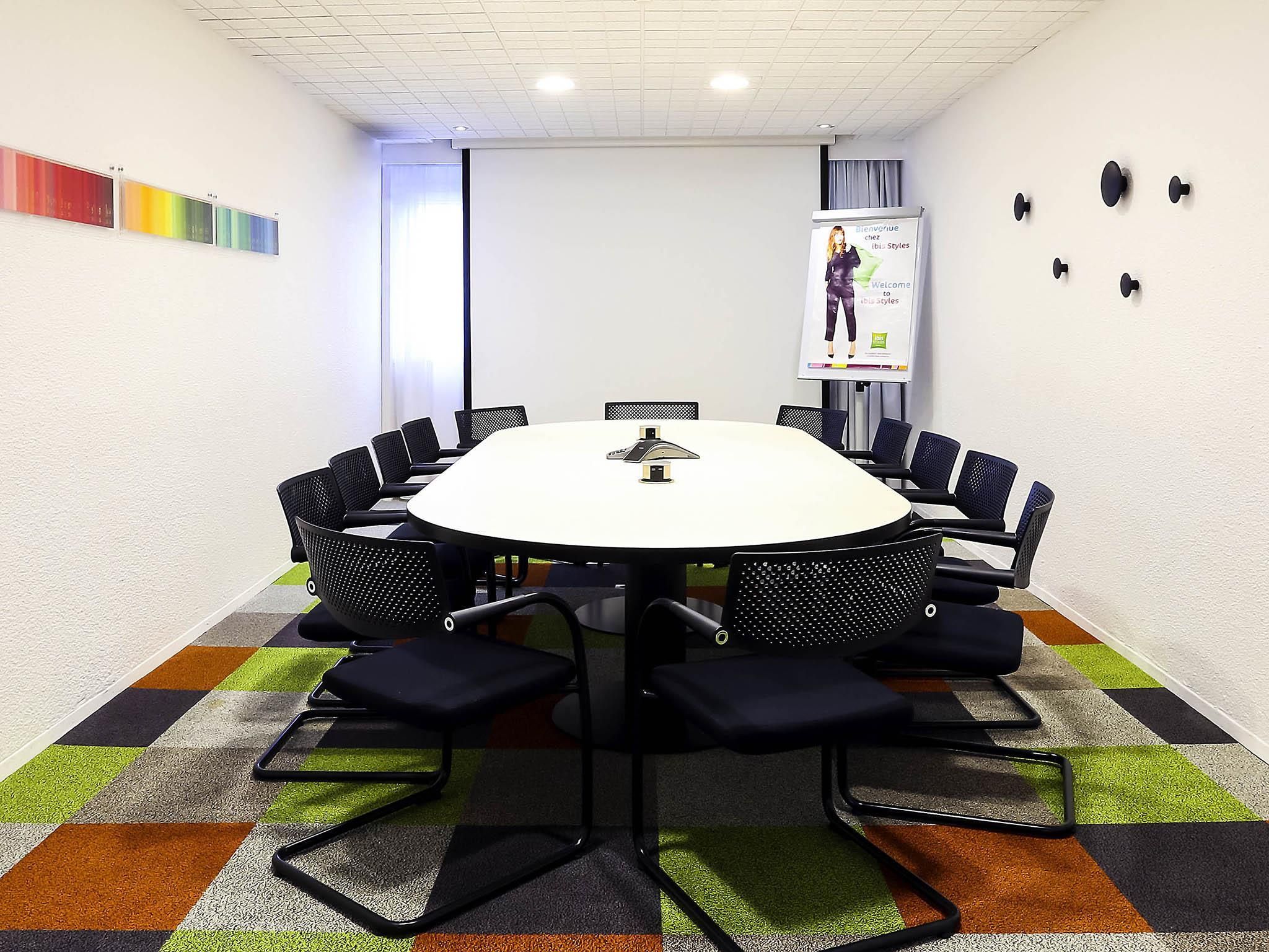 Une salle de conférence équipée d’une table ronde, de chaises noires, d’un écran de projection et d’une moquette colorée.