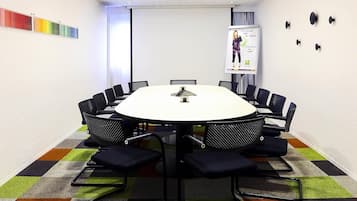 Uma sala de conferência com uma mesa redonda, cadeiras pretas, uma tela de projetor e um tapete colorido.