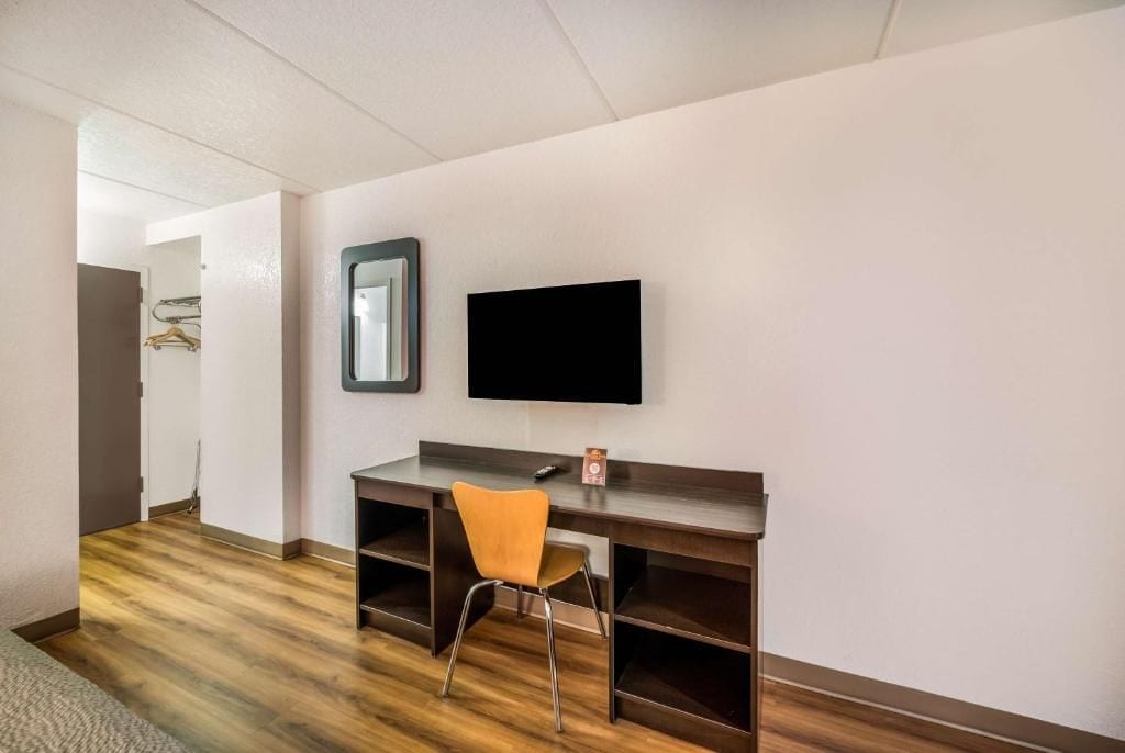 A hotel room with a wooden floor, a desk with a chair, a flat-screen TV mounted on the wall, and a door leading to another room.