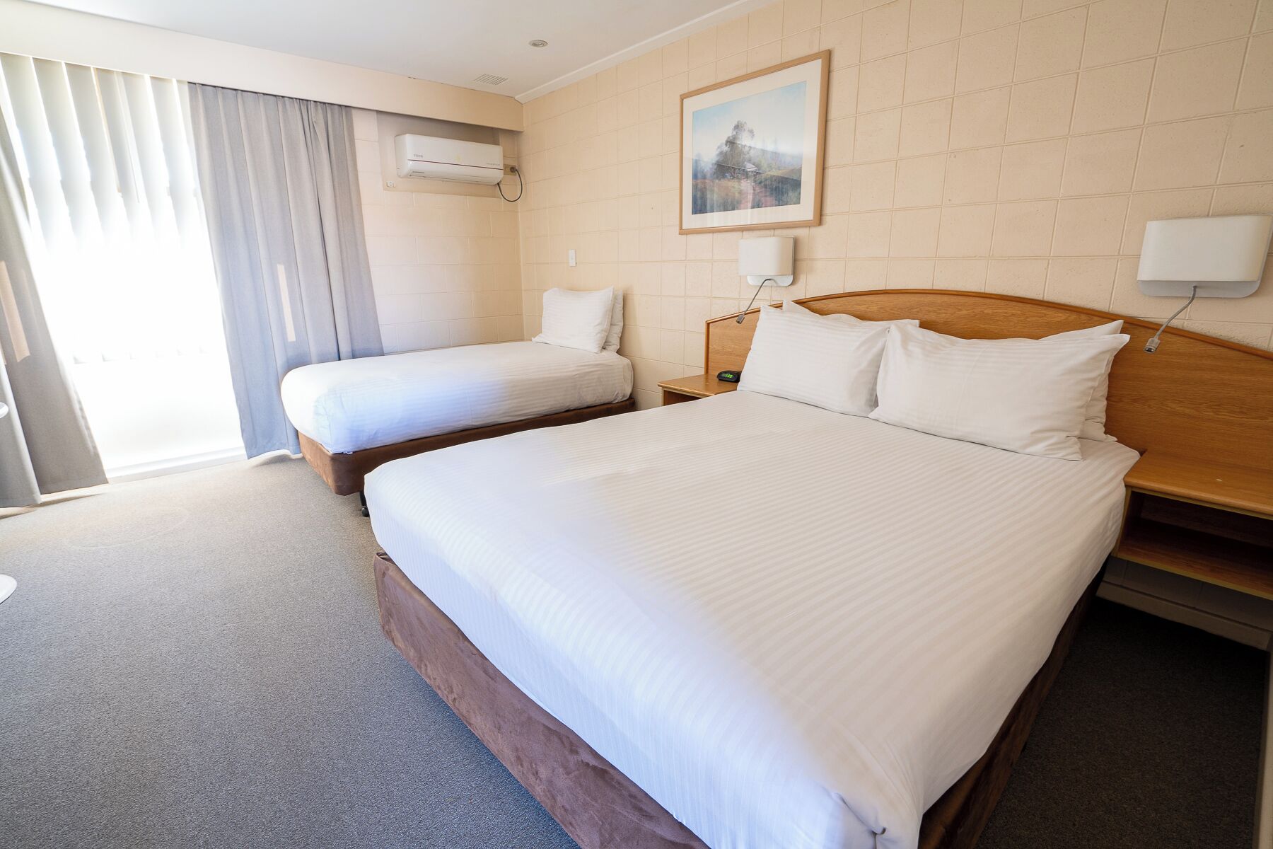 A hotel room with a large bed, a smaller bed, a nightstand, a lamp, an air conditioning unit, and a window with curtains.