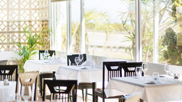 Een restaurant met gedekte tafels, grote ramen en tropische planten buiten.