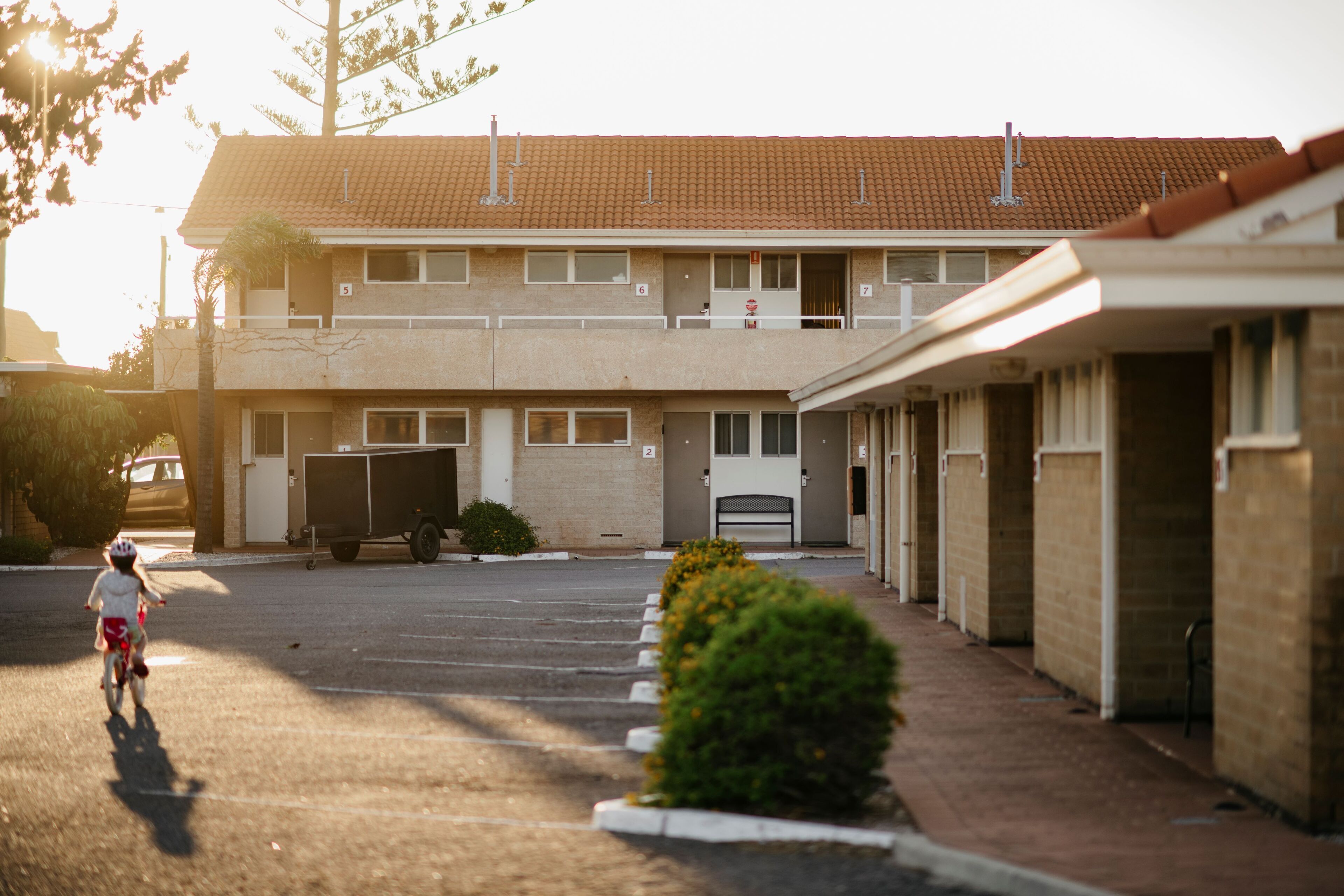 A two-story motel with a parking lot, a child riding a bicycle, and a row of bushes along the sidewalk.