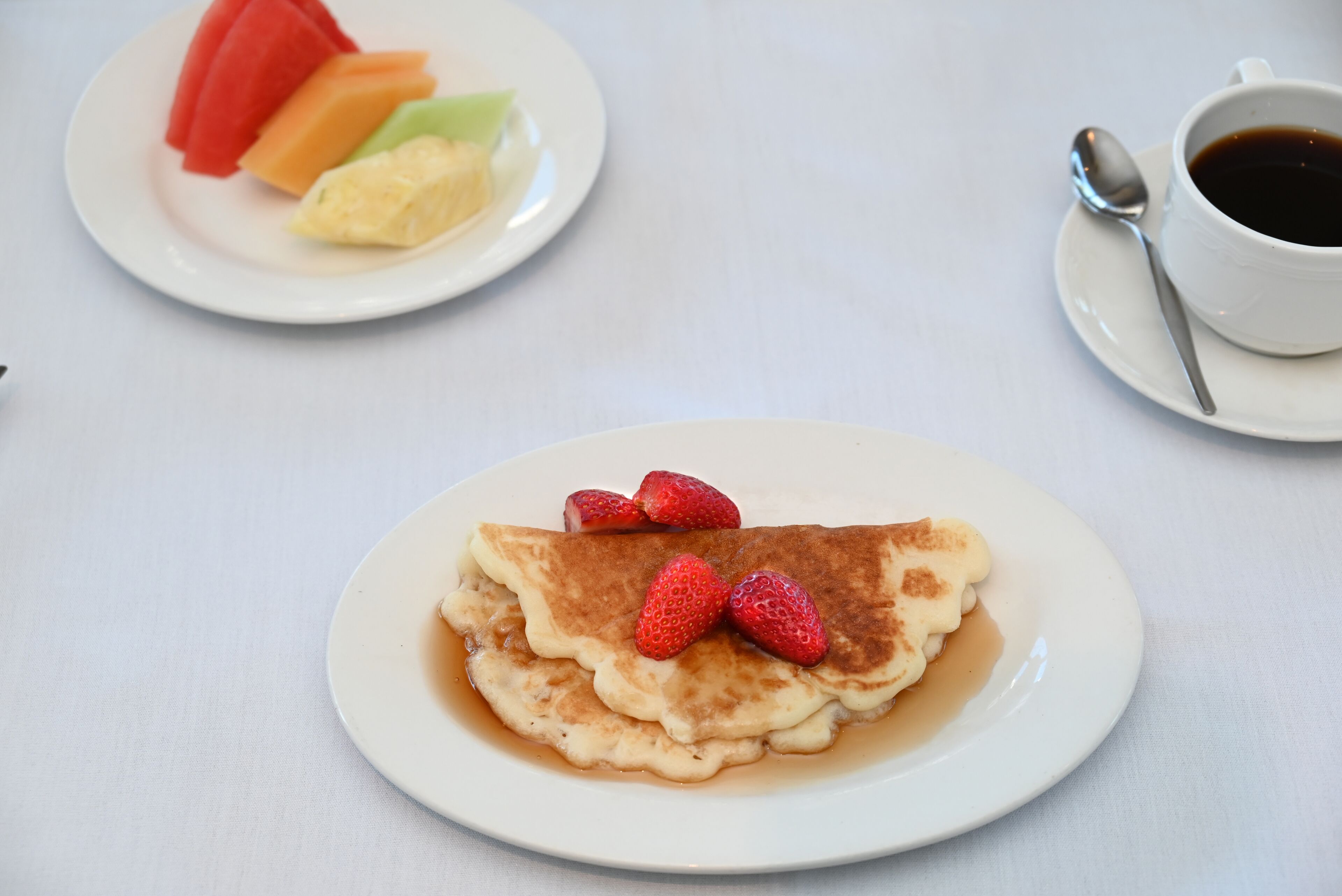A plate of pancakes with strawberries and syrup, a plate of fruit, and a cup of coffee with a spoon on a table.