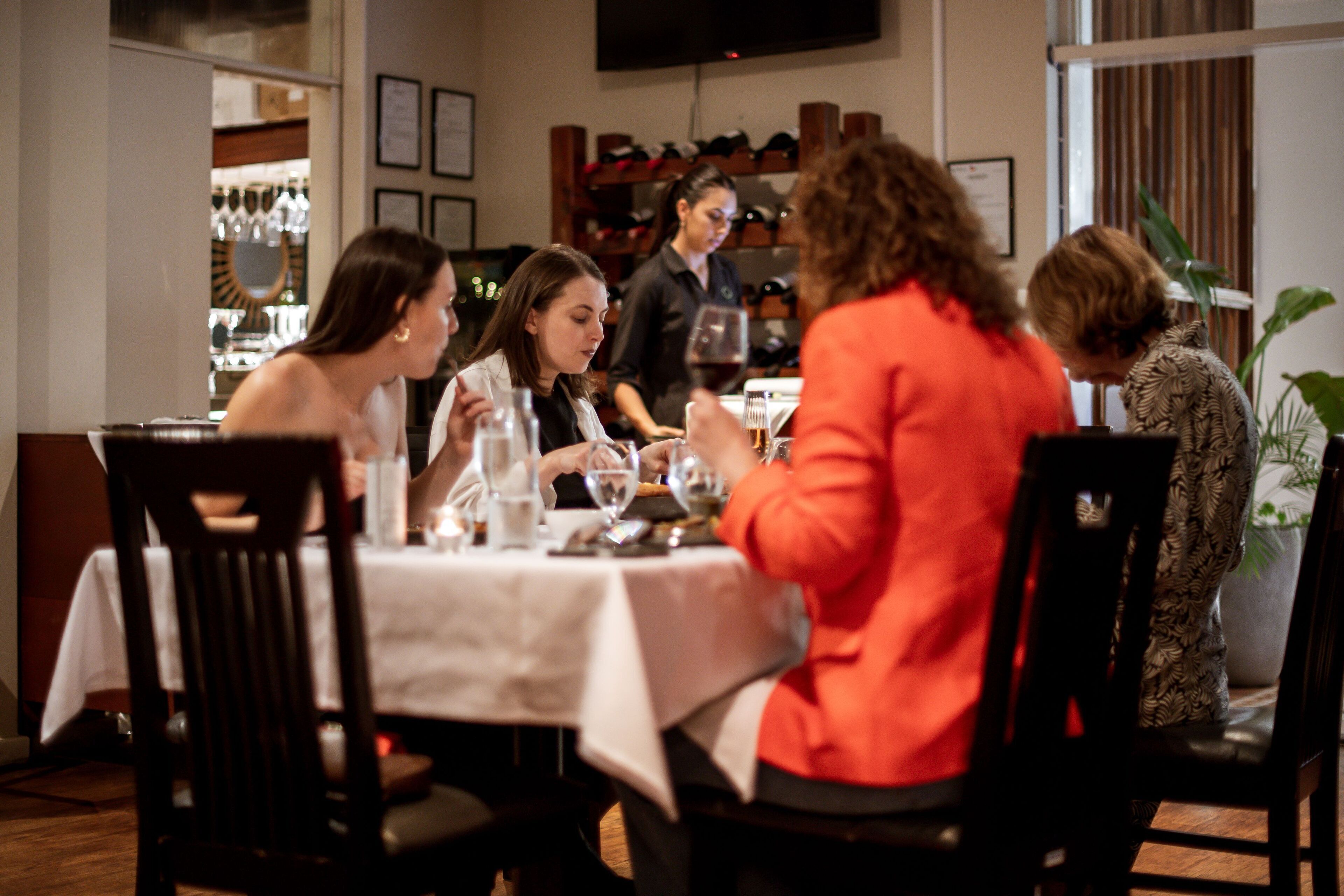 Un grupo de personas cenando en un restaurante con mantel blanco, copas de vino y un camarero al fondo.