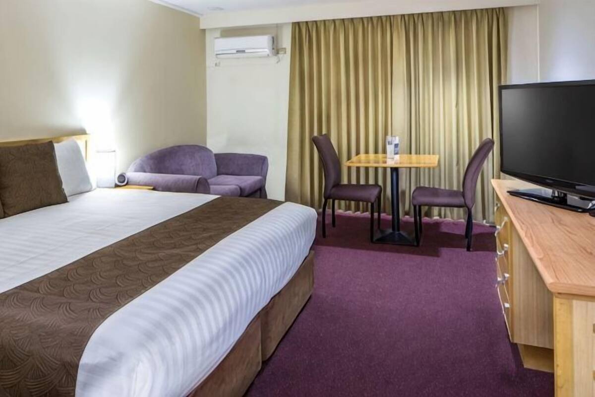 Et hotelværelse med en stor seng, et skrivebord med fjernsyn, to stole og et lille bord.