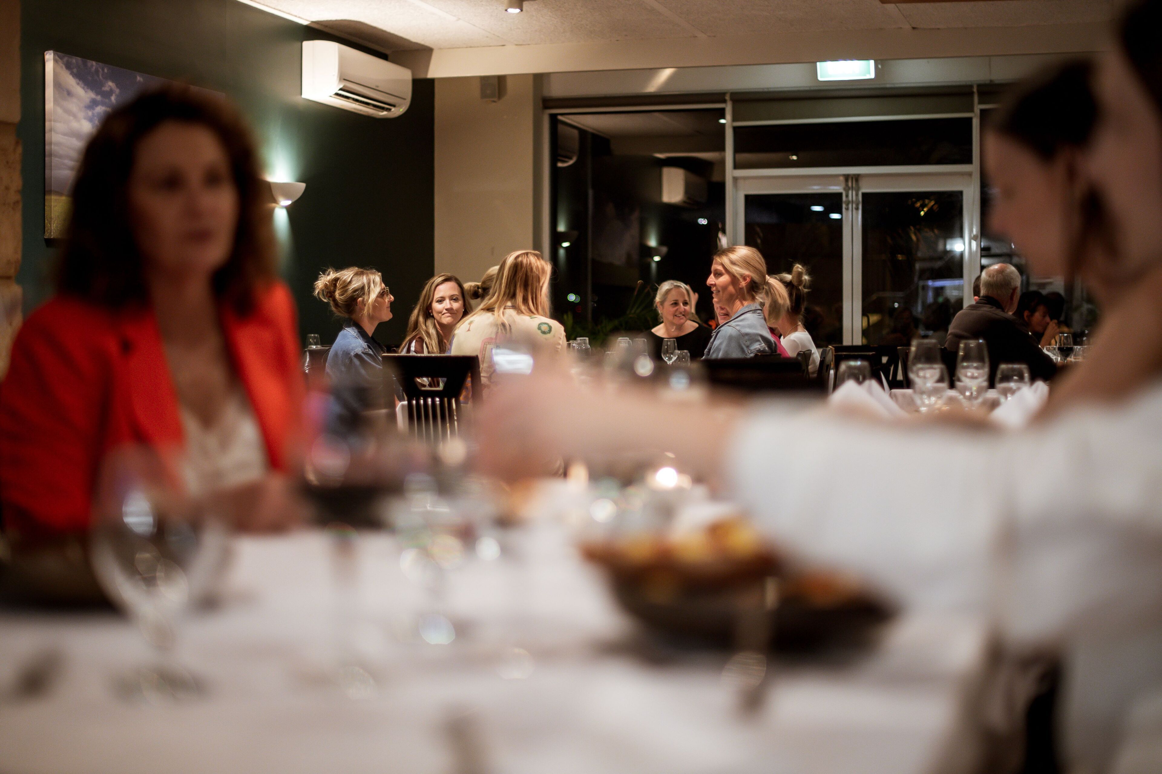 A restaurant with tables set for dining, a group of people engaged in conversation, and a view of the street outside.