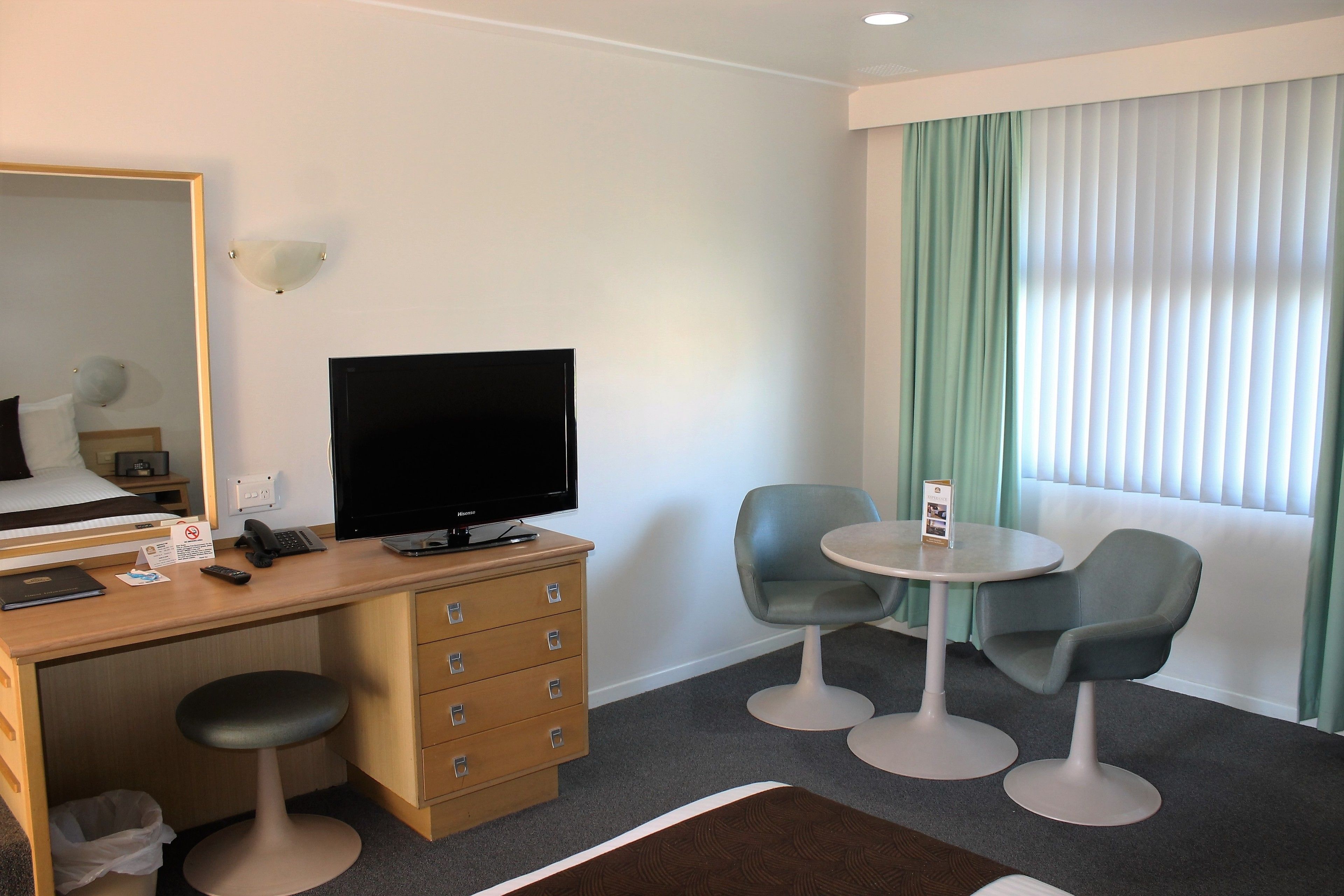 A hotel room with a wooden desk, a flat-screen TV, two chairs, a small table, a mirror, and a window with curtains.
