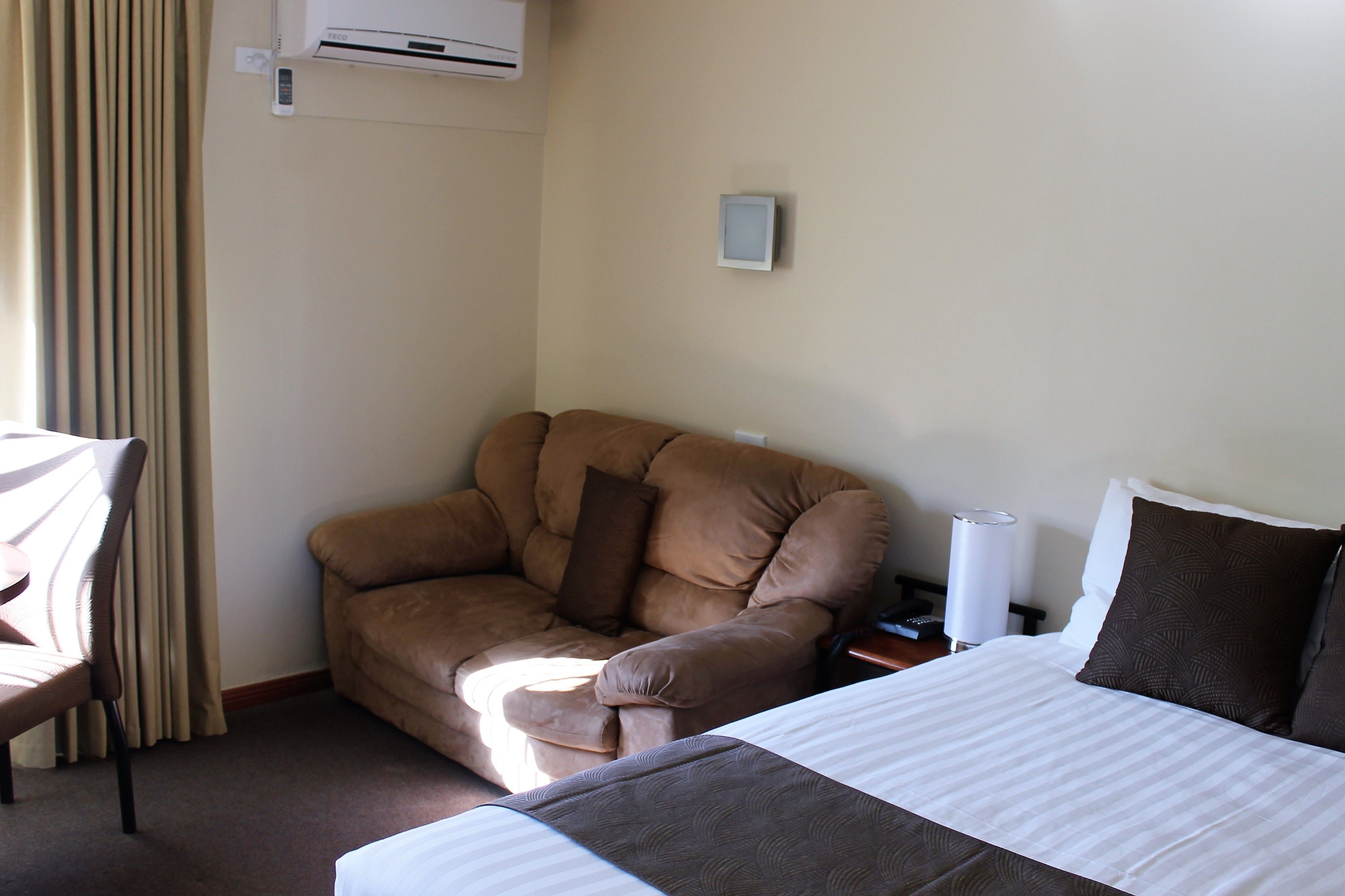 Una habitación de hotel con una cama, un sillón de cuero marrón, una silla, una mesita de noche con una lámpara y un equipo de aire acondicionado en la pared.