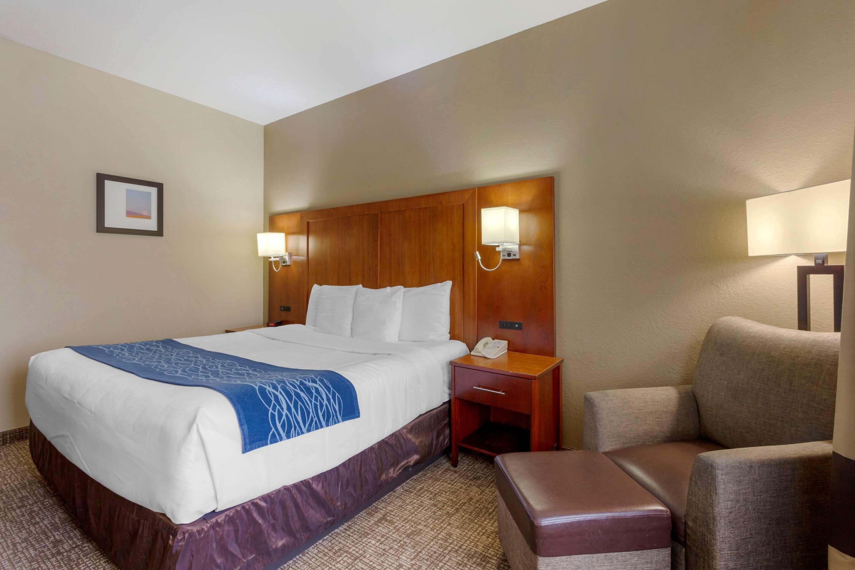 Quarto de hotel com cama grande, cabeceira de madeira, duas luminárias de cabeceira, mesinha, cadeira e um banquinho.