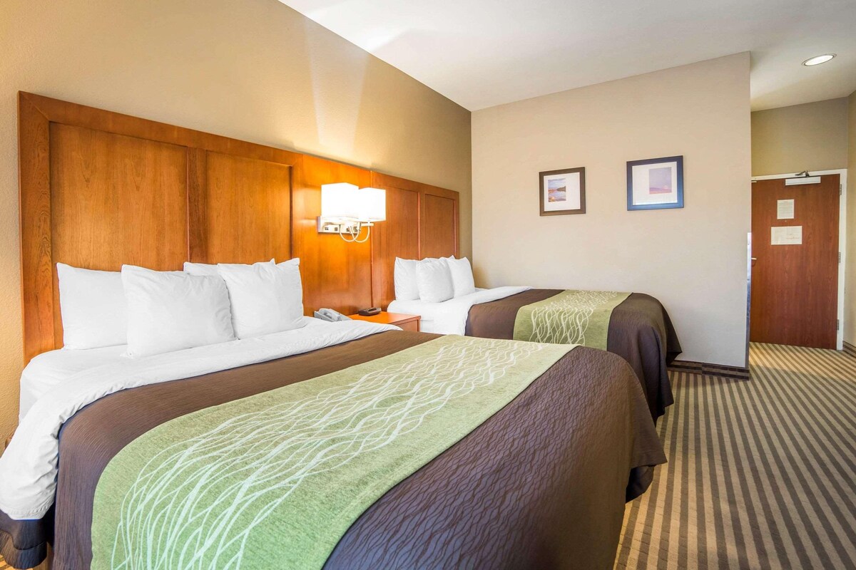 A hotel room with two beds, a wooden headboard, a bedside table with a lamp, and a framed picture on the wall.
