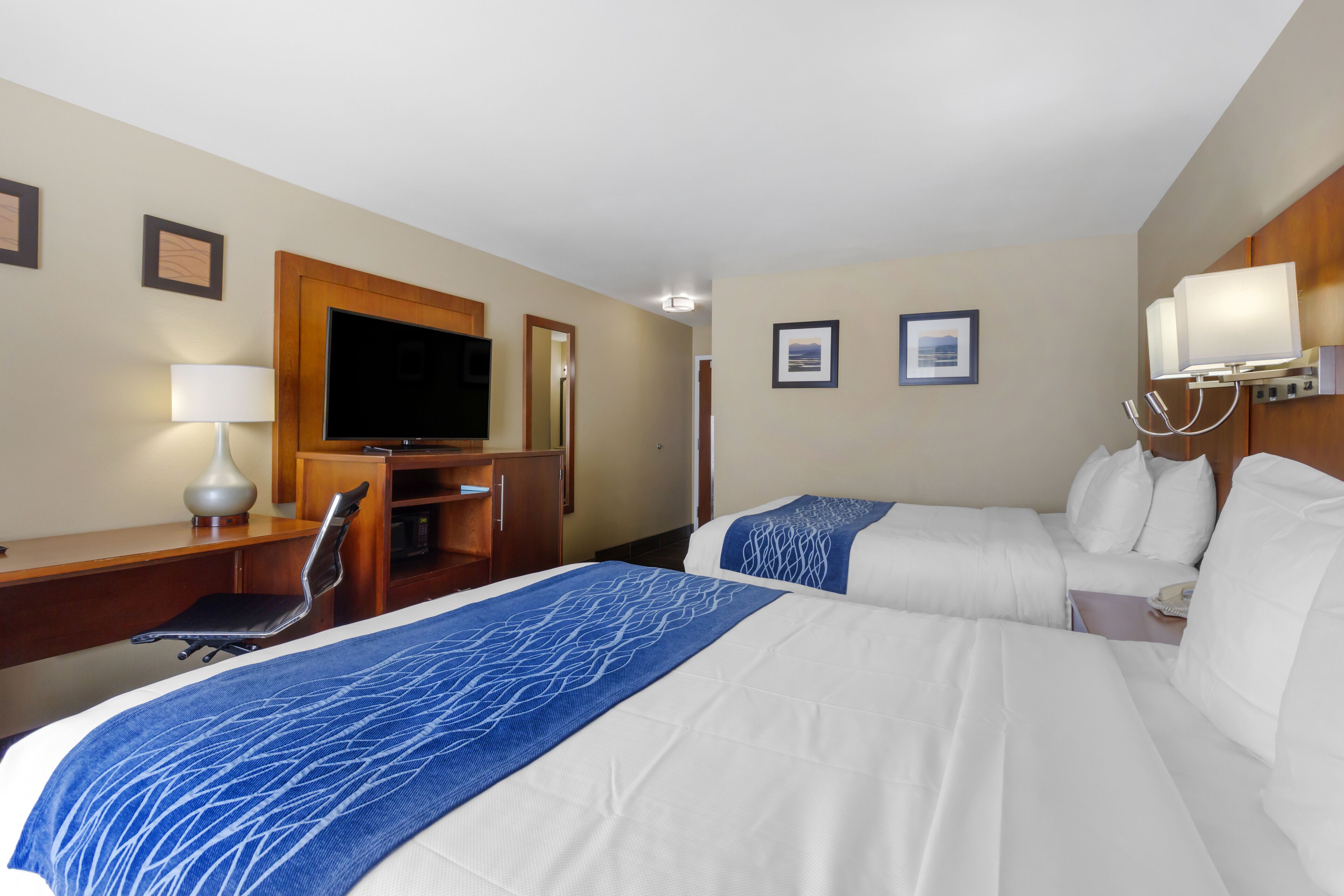 A hotel room with two beds, a desk with a chair, a television, and framed pictures on the walls.