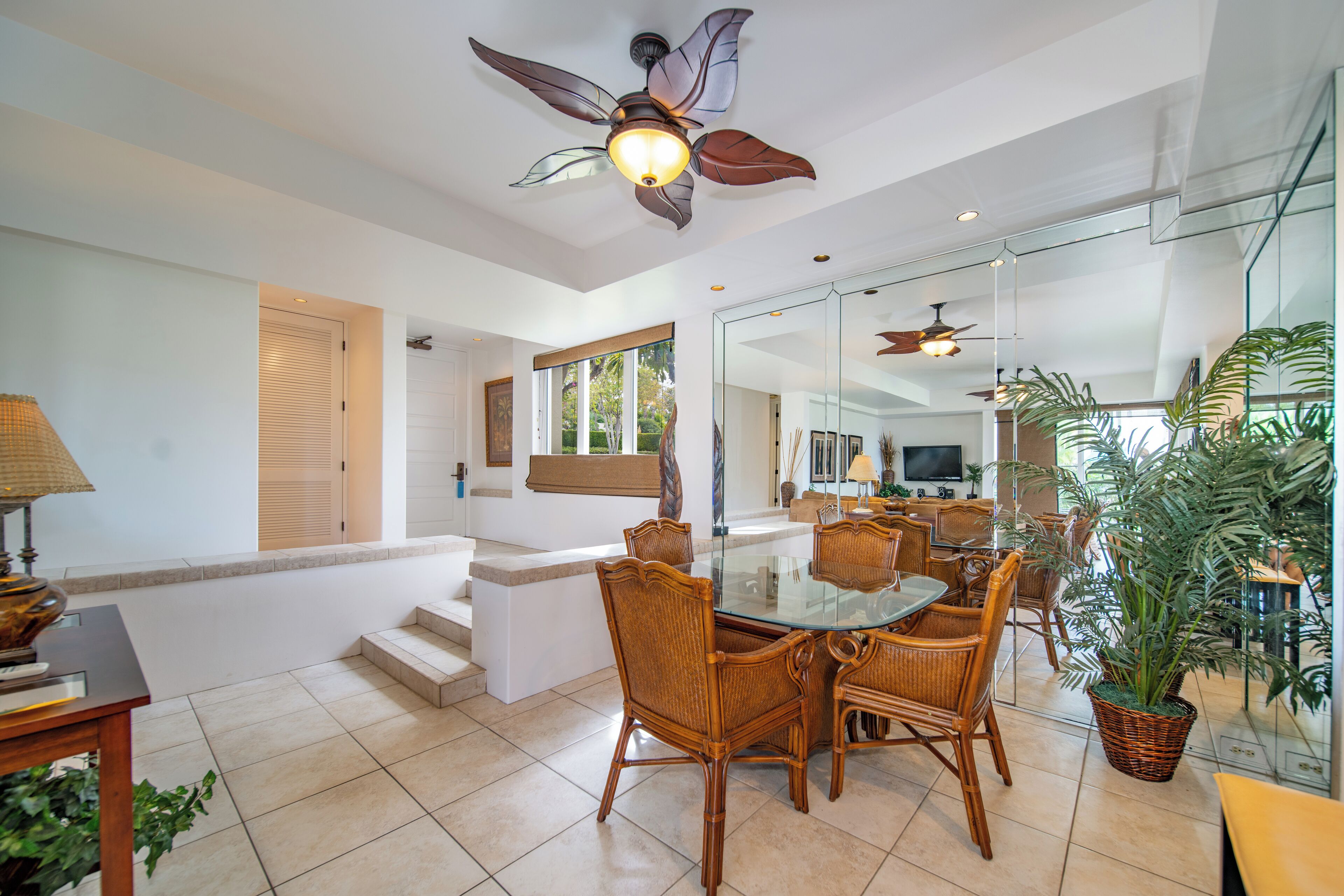 A dining area with a glass table and wicker chairs, a ceiling fan, and a large mirror reflecting the room.