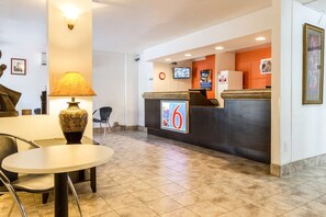 Zona de recepción de un hotel, con un mostrador, un reloj, un televisor y una mesa con sillas.