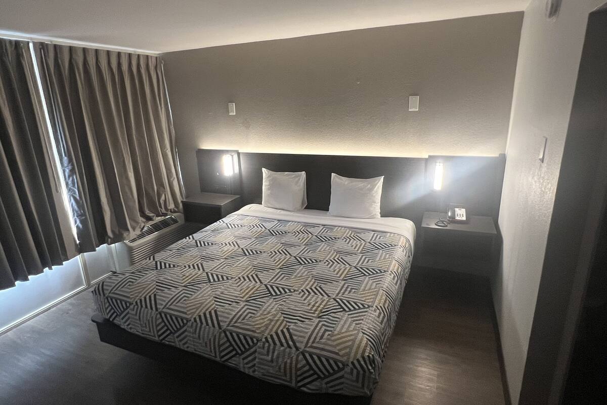 Hotellihuone, jossa on suuri sänky, kaksi yöpöytää, sängynpääty, jossa on sisäänrakennettu valaistus, ikkuna verhoilla ja seinälle asennettu termostaatti.