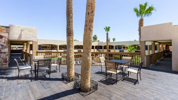 Un patio al aire libre con suelo de madera, sillas y mesas de metal, rodeado de palmeras y un edificio con fachada color beige.