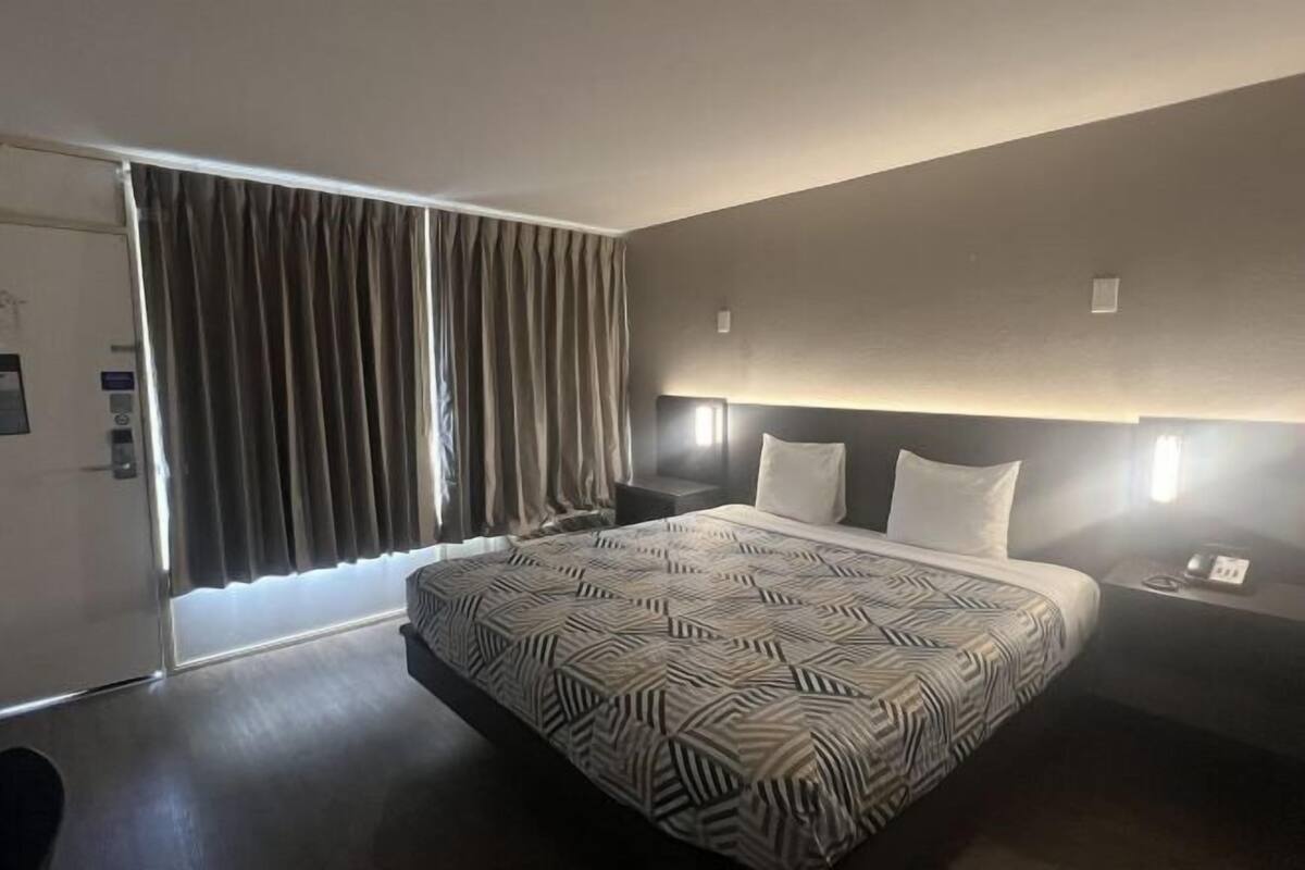 Moderni hotellihuone, jossa on suuri sänky, yöpöytä, yövalaisin, ikkuna verhoilla ja sängynpääty, jossa on sisäänrakennettu valaistus.