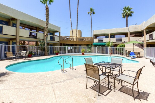 A swimming pool with lounge chairs and tables, surrounded by palm trees and a multi-story building with balconies.