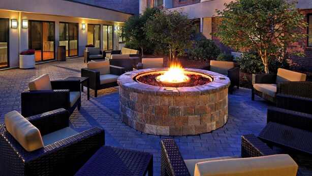 A circular fire pit surrounded by wicker furniture in an outdoor patio area with brick flooring and a brick building in the background. - Courtyard by Marriott Hartford Cromwell (Cromwell)
