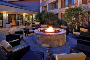 A circular fire pit surrounded by wicker furniture in an outdoor patio area with brick flooring and a brick building in the background. - Courtyard by Marriott Hartford Cromwell (Cromwell)