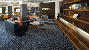 A modern lobby with a dark blue patterned carpet, a black sofa with orange cushions, a round glass coffee table, and a wooden bookshelf with decorative items. - Courtyard by Marriott Hartford Cromwell (Cromwell)