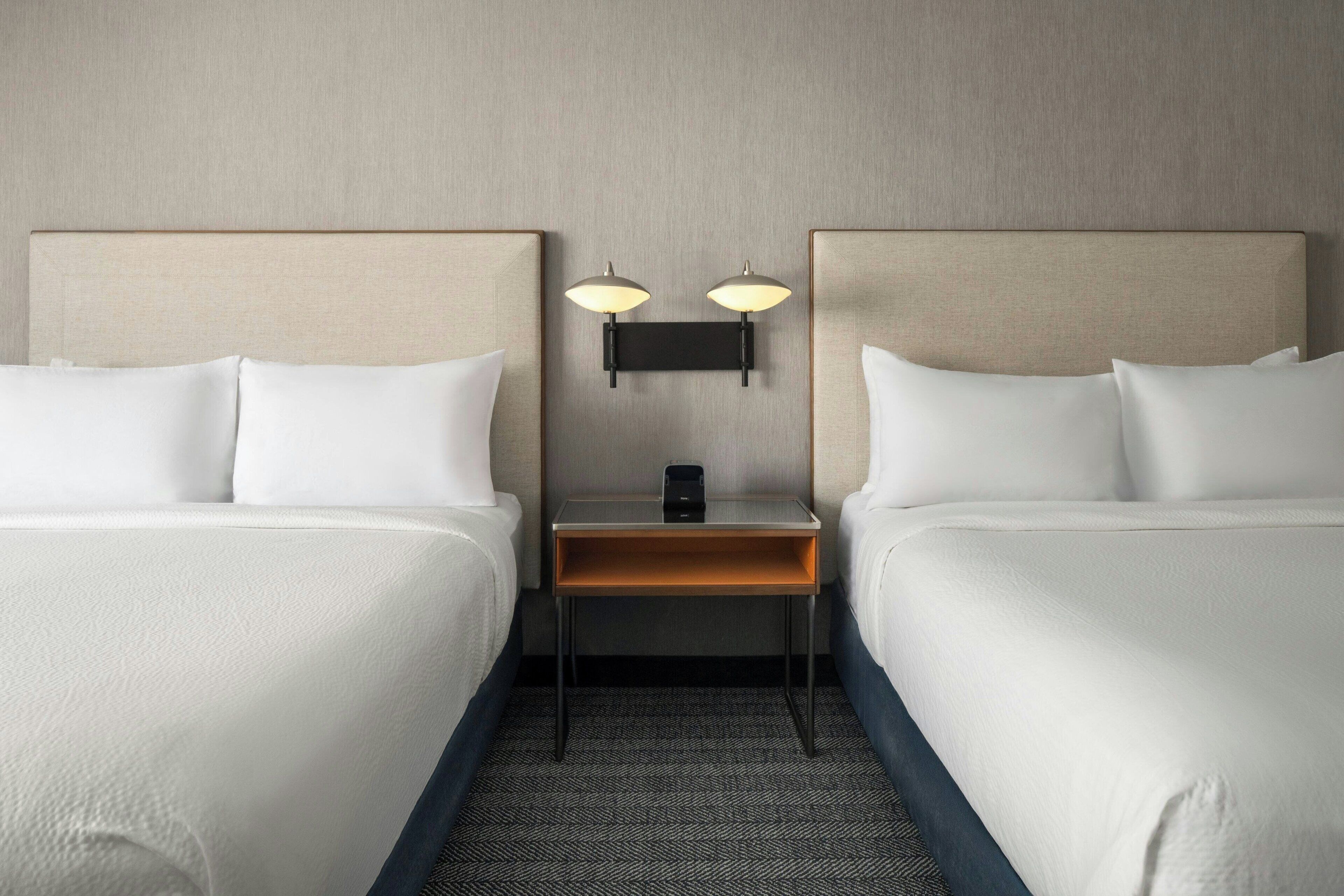 A hotel room with two beds, a nightstand with a lamp, and a small table.