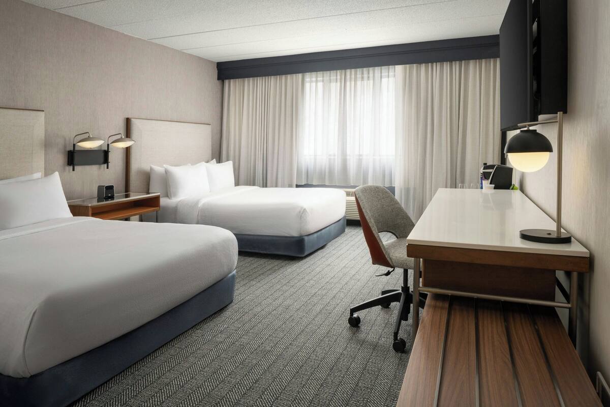 A hotel room with two beds, a desk, a chair, a TV, and a window with curtains.