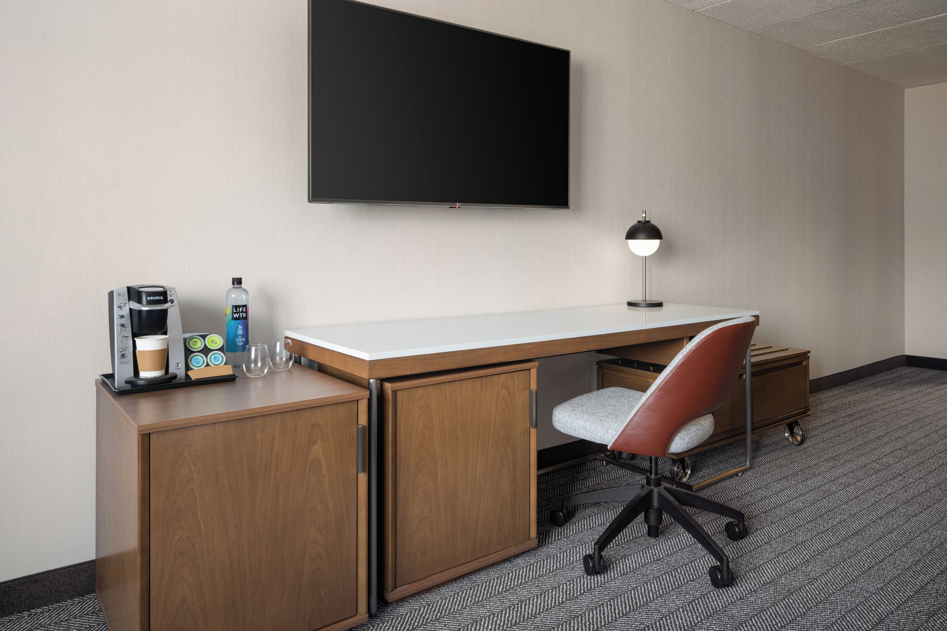 A hotel room with a wooden desk, a chair, a flat-screen TV, a coffee maker, a bottle of water, and a glass.