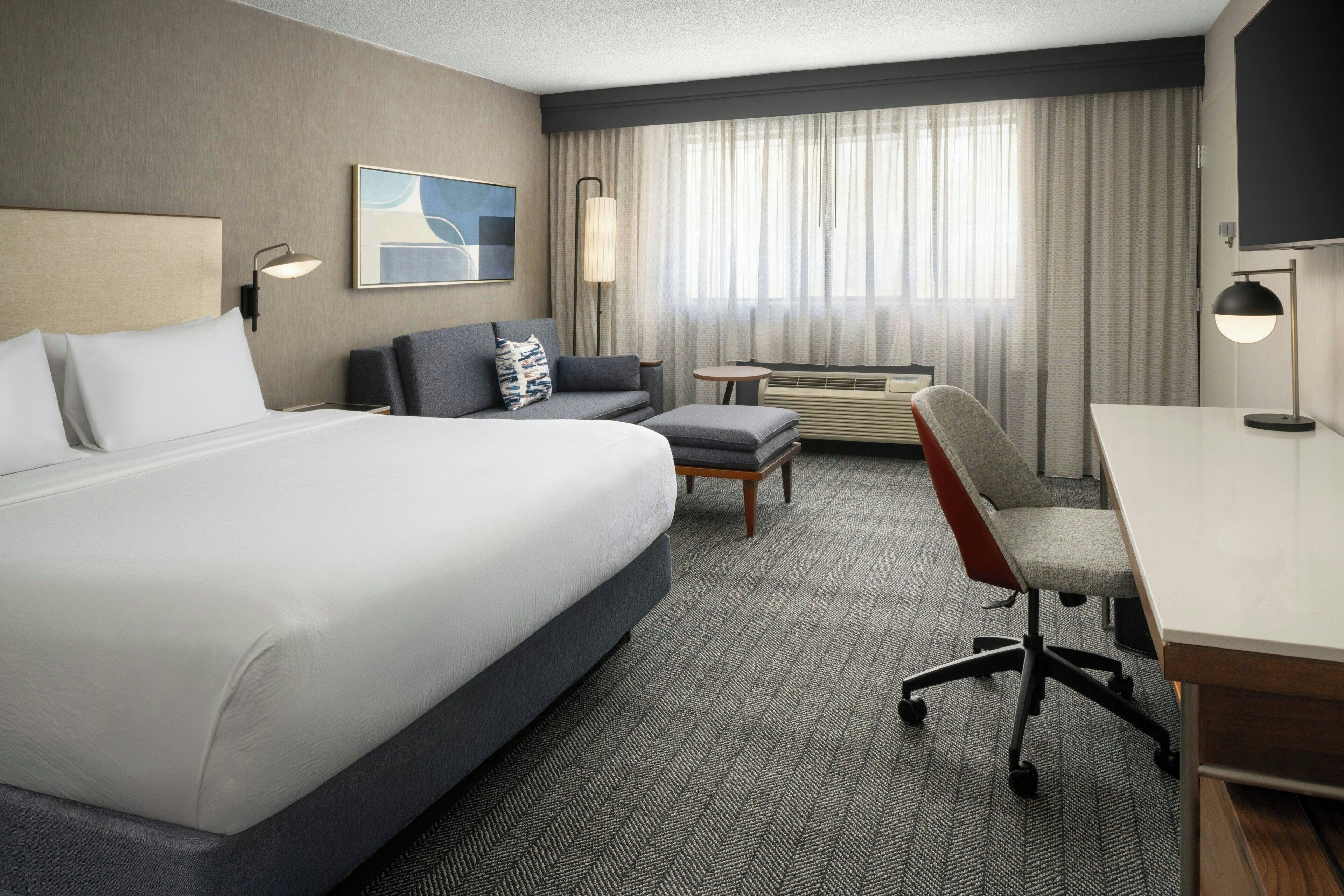 A hotel room with a large bed, a desk, a chair, a small table, and a sofa.
