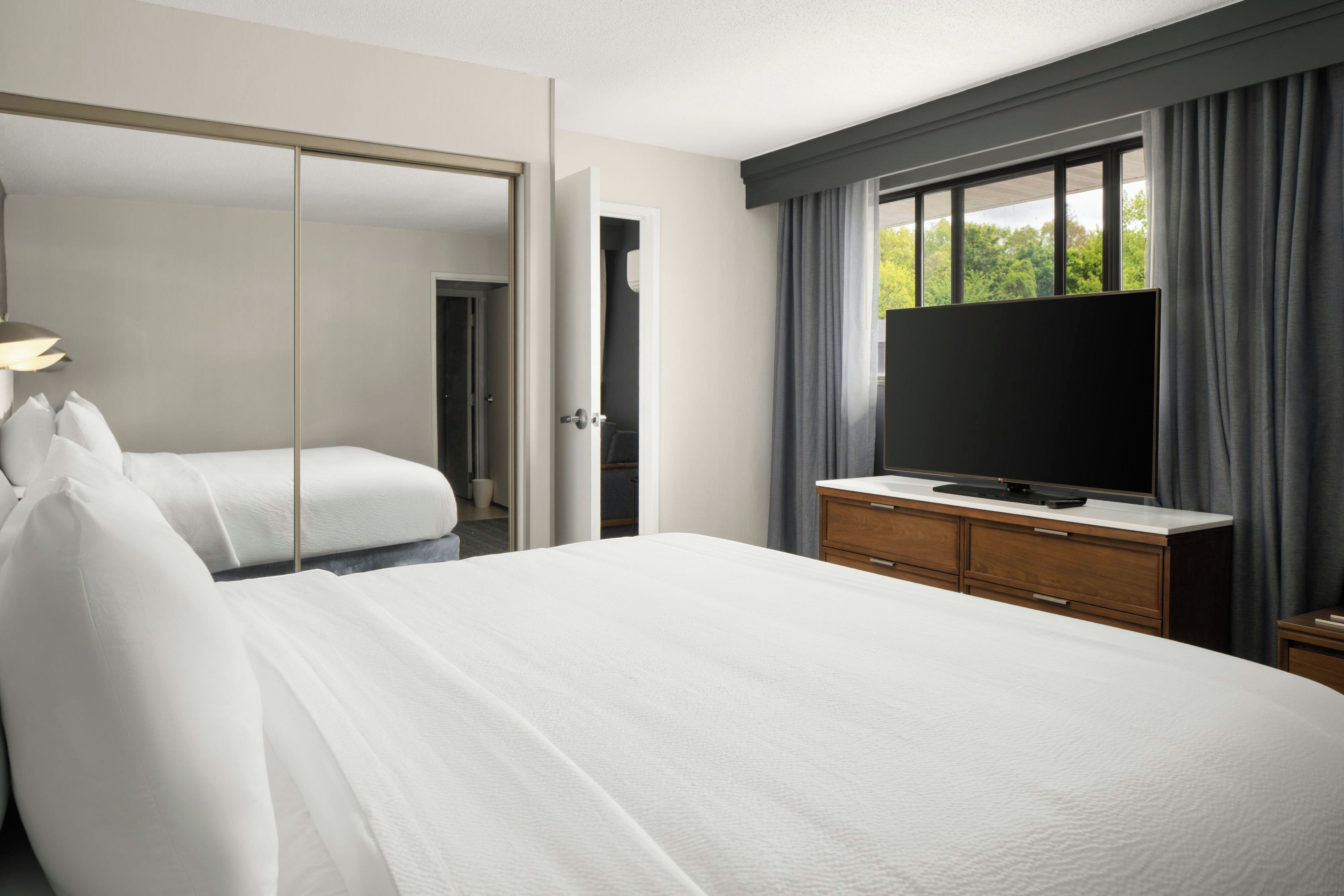 A hotel room with a bed, a television on a dresser, a mirror, and a window with curtains.