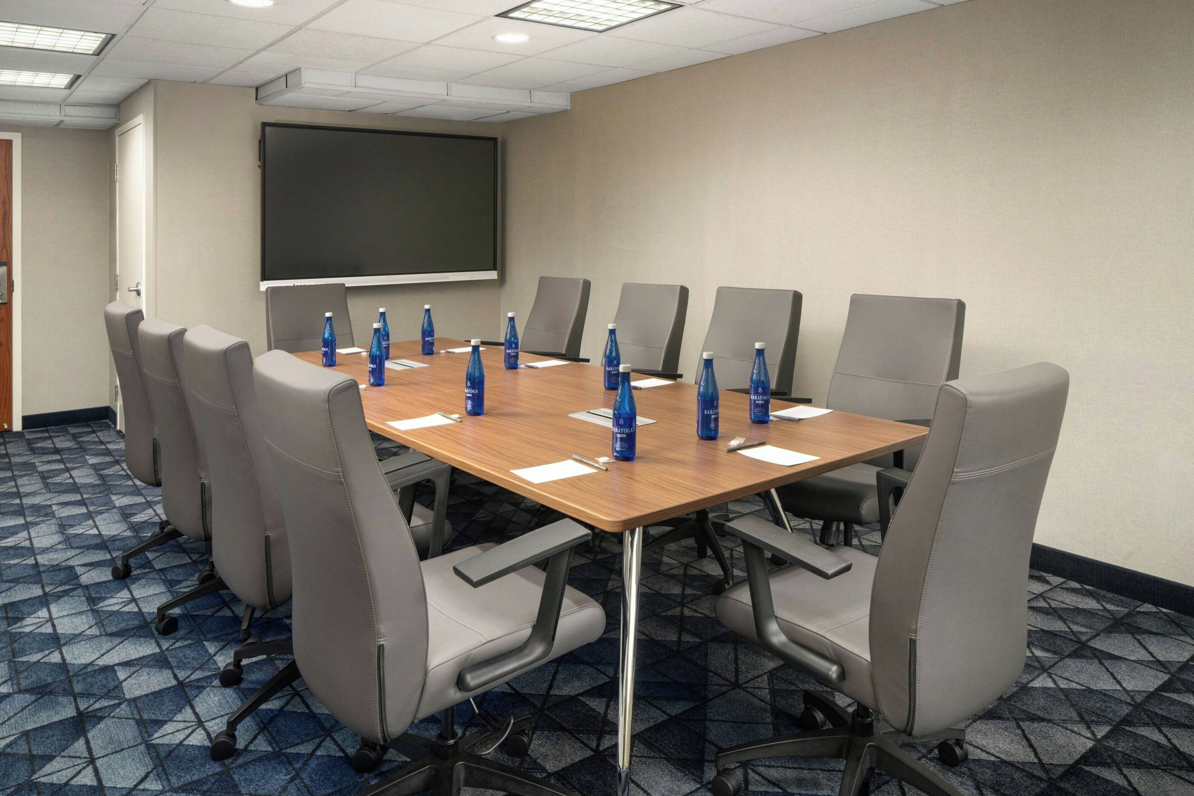 A conference room with a large table, eight chairs, a projector screen, and water bottles on the table.