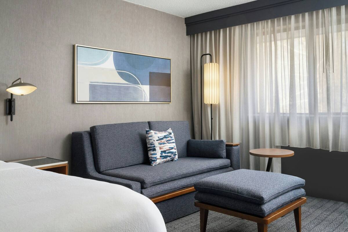 A hotel room with a grey sofa, a small round table, a lamp, and a painting on the wall.