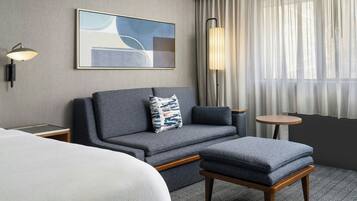 A hotel room with a grey sofa, a small round table, a lamp, and a painting on the wall.