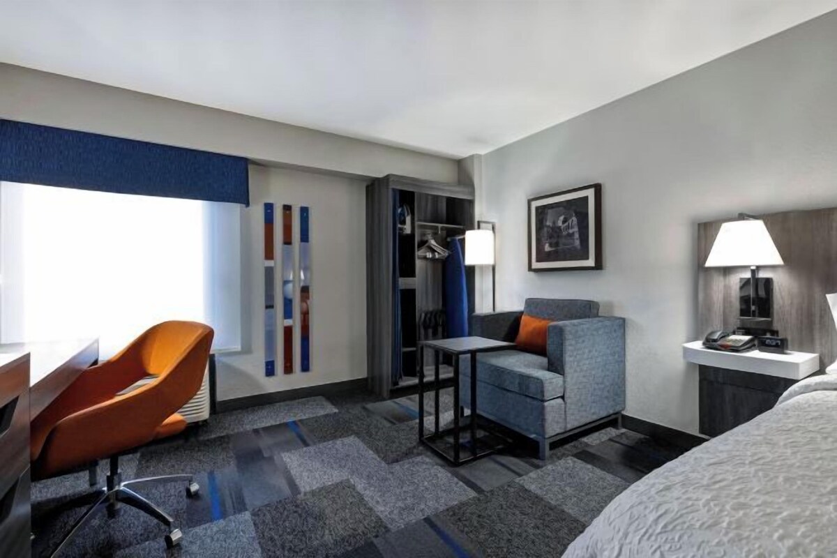 A hotel room with a bed, a desk with a chair, a small table, a wardrobe, and a nightstand with a lamp.