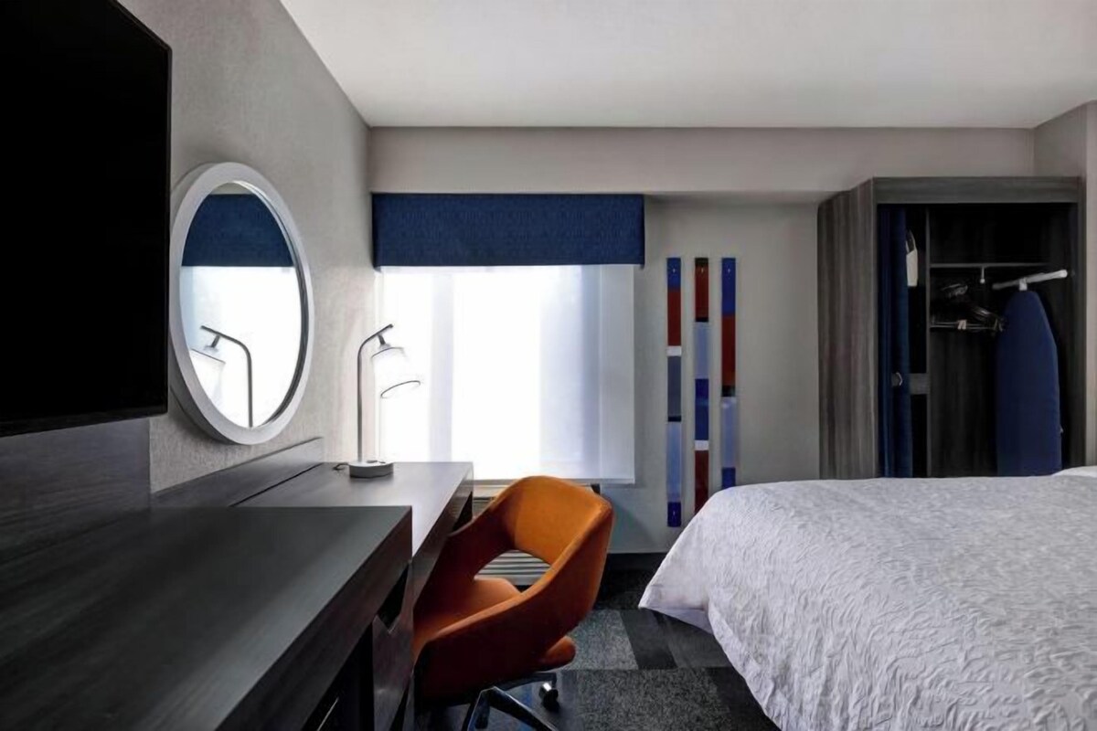 A hotel room with a bed, a desk with a chair, a mirror, and a wardrobe.