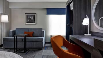 A hotel room with a grey sofa, a small table, an orange chair, a desk with a lamp, a framed picture on the wall, and a window with a blue curtain.