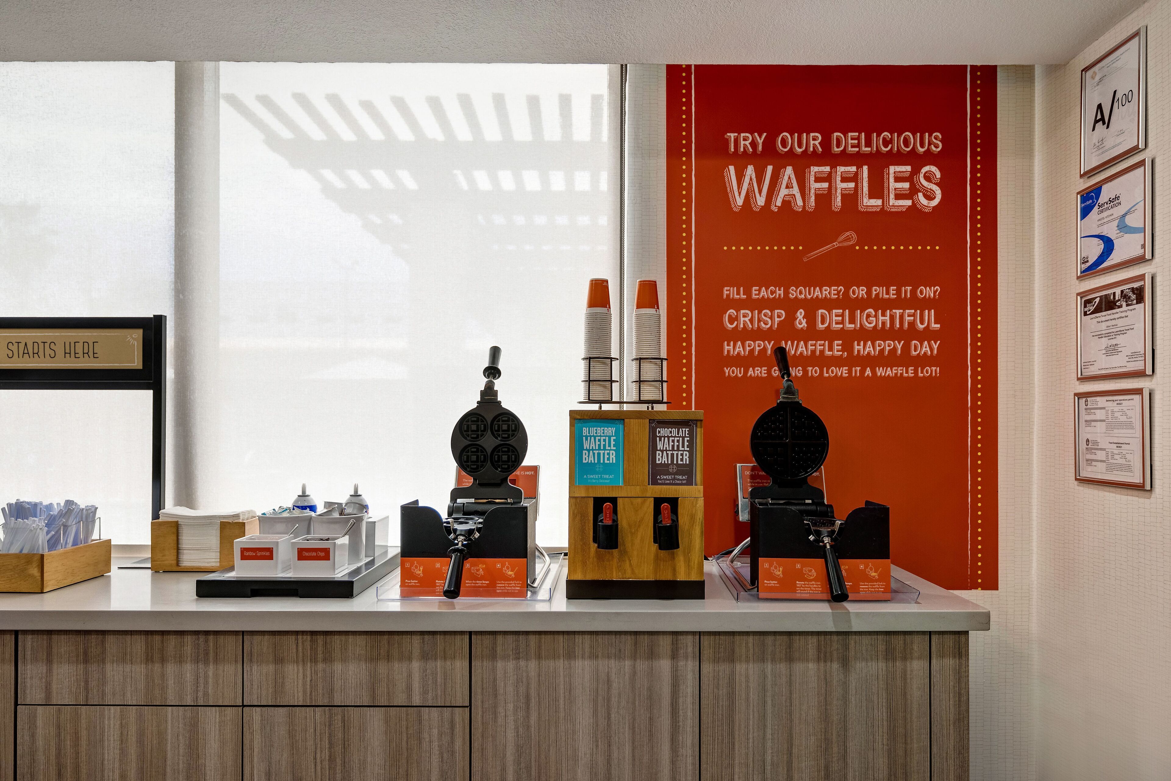Una estación de gofres con máquinas para hacer gofres y dispensadores de masa, un letrero que promociona los gofres y un letrero que dice 'Aquí Comienza'.