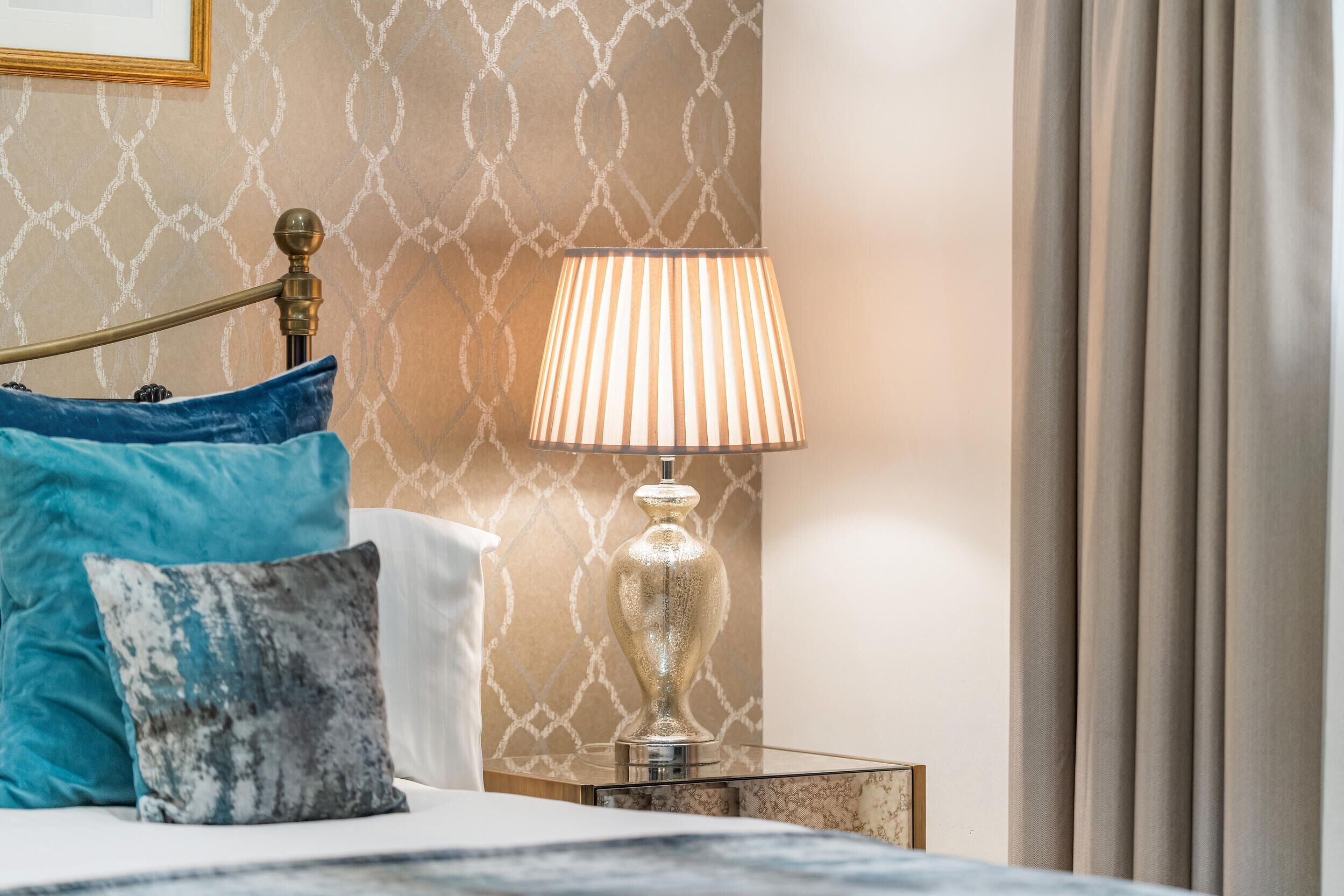 A neatly made bed with blue and grey pillows, a brass bed frame, and a bedside table with a lamp.