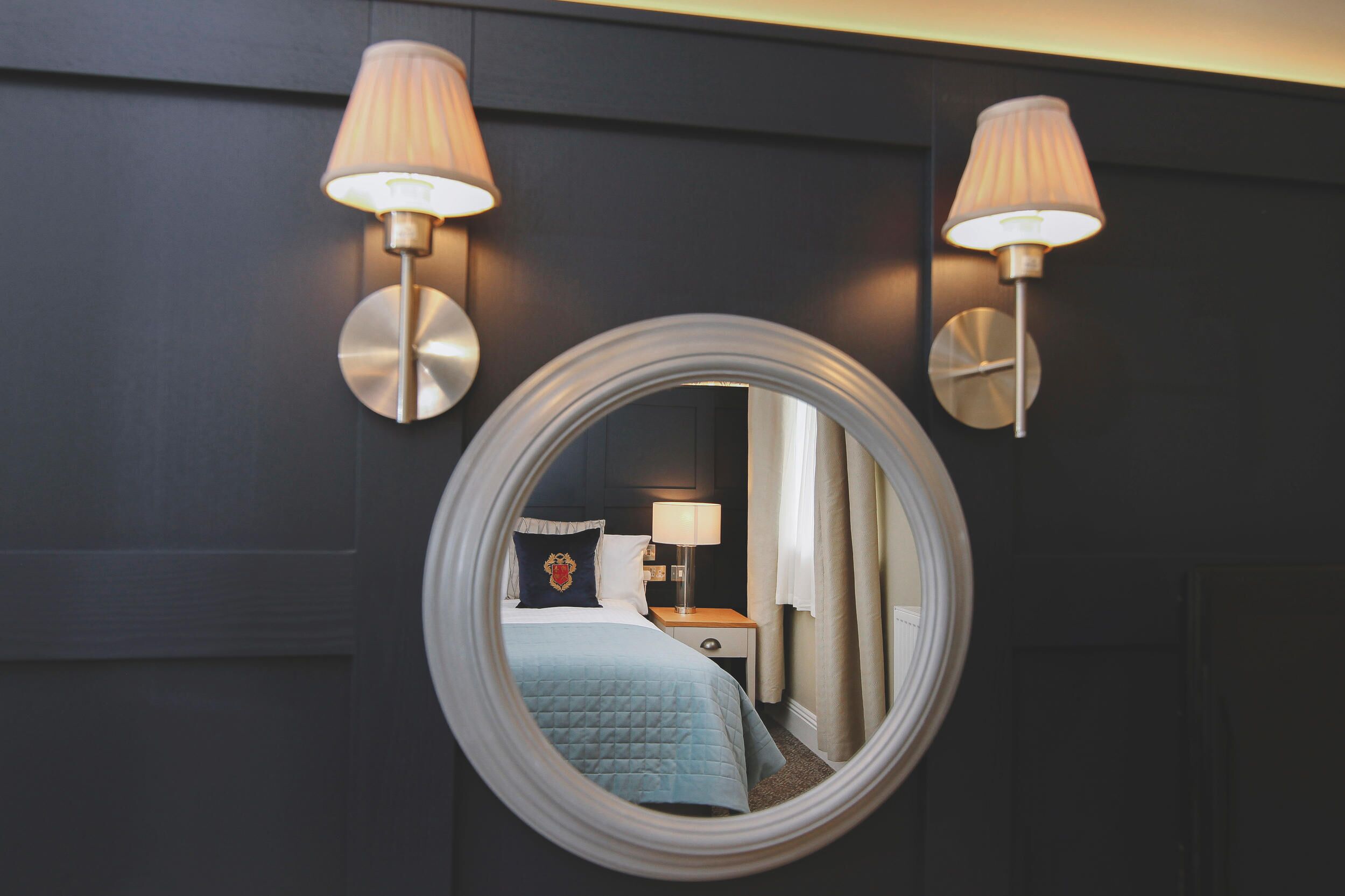 Une chambre d’hôtel avec un miroir rond, deux lampes fixées au mur, un lit recouvert d’une courtepointe bleue, une table de chevet et une commode.