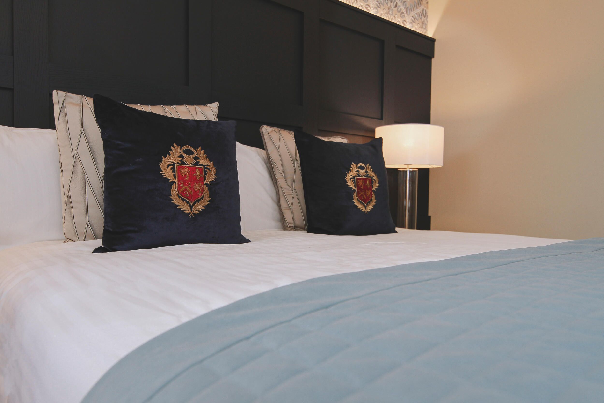 A neatly made bed with white linens and two dark pillows with gold emblems.