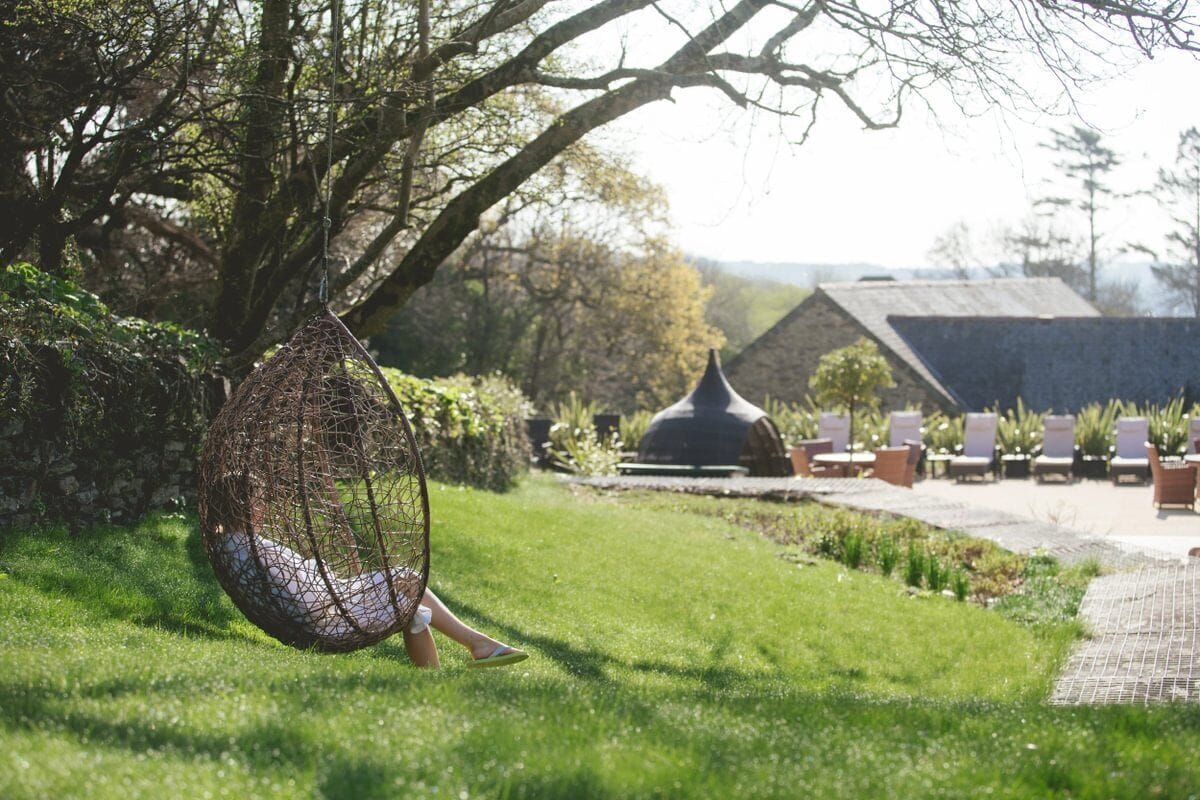 Een hangmat in een weelderige tuin met een prieel en lounge fauteuils op de achtergrond.